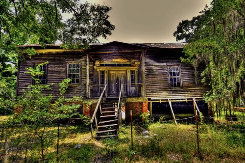 Abandoned wooden house with a front porch, overgrown vegetation, and a weathered facade under a bright sky.