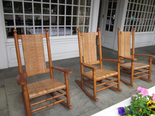 Three wooden rocking chairs with woven seats, placed on a stone patio near a windowed building. Colorful flowers in the foreground.