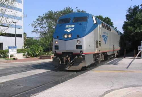 An Amtrak train approaches a crossing, surrounded by greenery and buildings in the background.