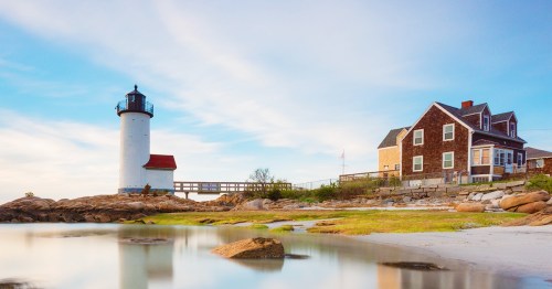 A scenic lighthouse beside a coastal house, with calm waters and a clear sky in the background.
