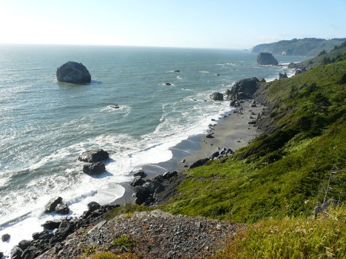 A scenic coastal view with rocky cliffs, waves crashing on the shore, and distant hills under a clear blue sky.