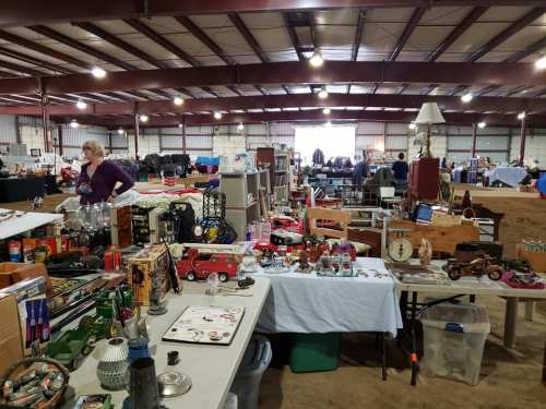 A bustling indoor market filled with various antiques and collectibles on tables, with shoppers browsing in the background.