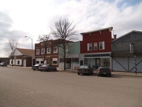 A small town street featuring several storefronts, parked cars, and a cloudy sky.