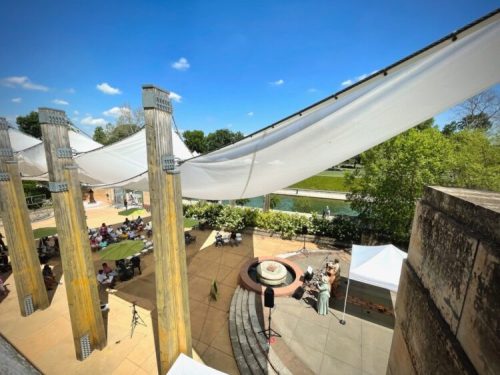 A sunny outdoor venue with a large canopy, seating area, and a small stage, surrounded by greenery and a water feature.