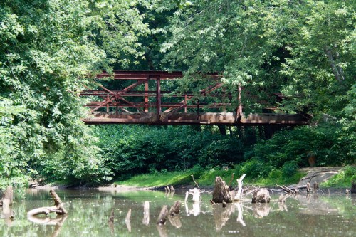 A rusted metal bridge spans a calm, reflective pond surrounded by lush green trees and fallen logs.