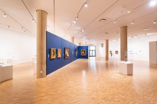 A spacious art gallery featuring paintings on blue walls and sculptures displayed on white pedestals.