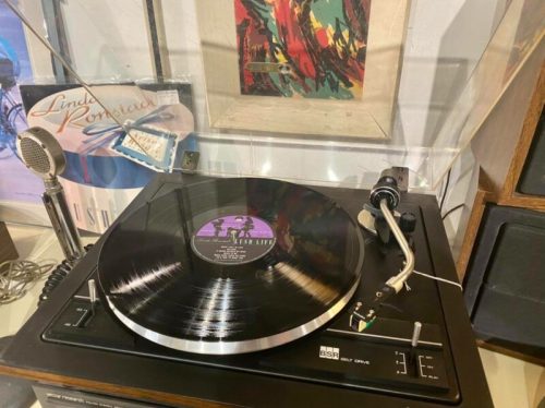 A vintage turntable playing a vinyl record, with framed artwork and a microphone in the background.