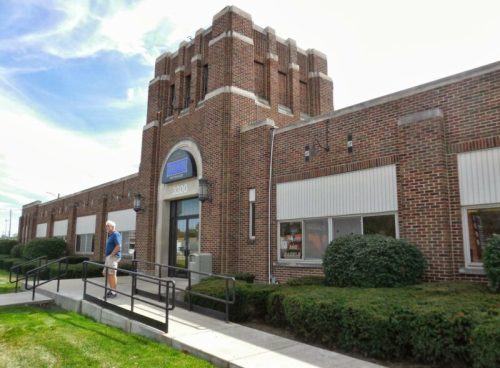 A man walks towards a brick building with a decorative tower and large windows, surrounded by greenery.