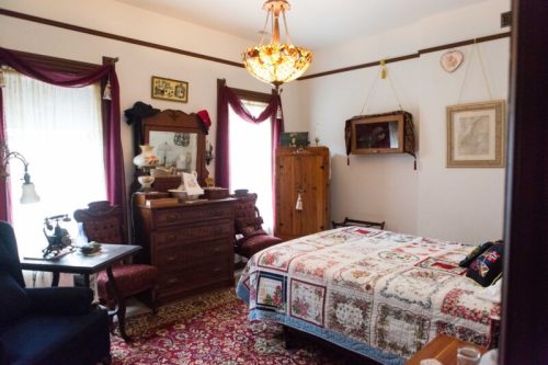 A cozy bedroom featuring a quilted bed, vintage furniture, and warm lighting from a decorative chandelier.
