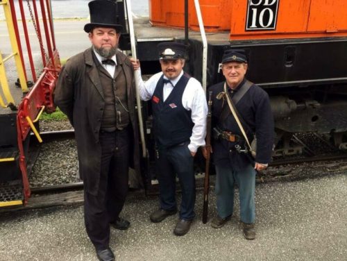 Three men in historical attire stand by a train, with one in a top hat, one in a conductor's uniform, and one in a military outfit.