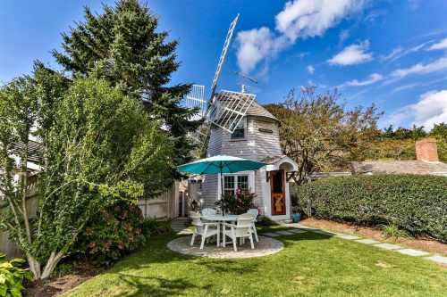 A quaint house with a windmill, surrounded by greenery and a patio set under an umbrella on a sunny day.