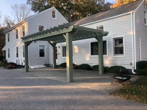 A white house with a green pergola in the driveway, surrounded by shrubs and a gravel path.