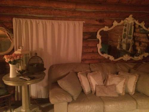 Cozy cabin interior featuring a beige couch, decorative pillows, a mirror, and a curtain against a log wall.