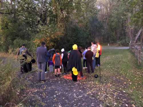 A group of people in a wooded area, some in costumes, gathered around a small fire during dusk.