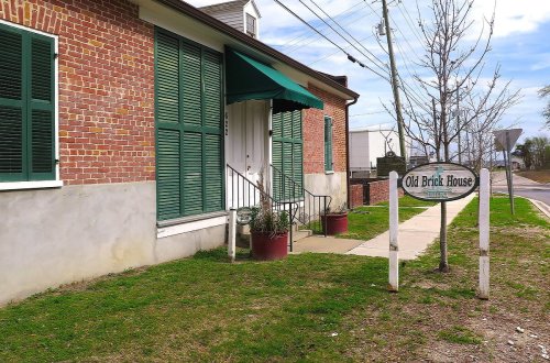 A brick building with green shutters and an awning, featuring a sign that reads "Old Brick House" near a sidewalk.