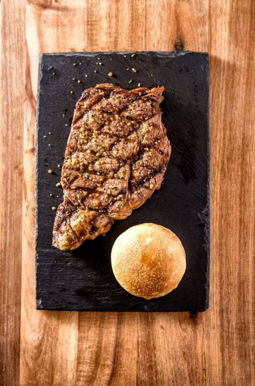 A grilled steak with seasoning served on a black slate plate alongside a small bread roll, on a wooden table.