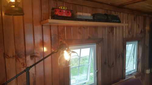 A wooden shelf with toy trains sits above two windows in a cozy, wooden-paneled room. A lamp is nearby.