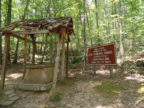 A wooden well house in a forest, with a sign about its history and construction by John Mauldin in the late 1800s.