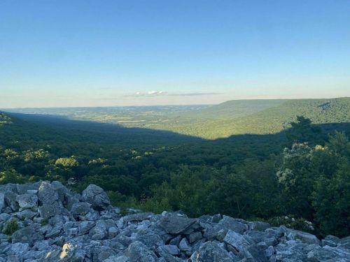 A panoramic view of a lush green valley surrounded by mountains under a clear blue sky. Rocky foreground.