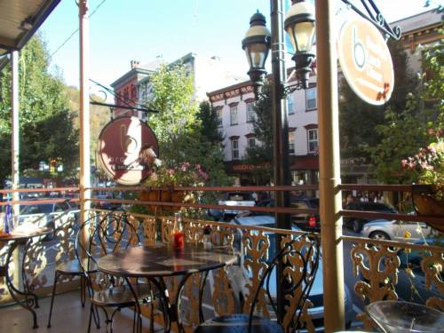 A cozy café balcony with tables, overlooking a street lined with trees and historic buildings.