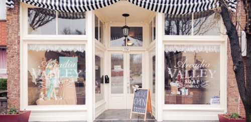 A charming soap shop with large windows, a striped awning, and a sign reading "Now Open" outside.
