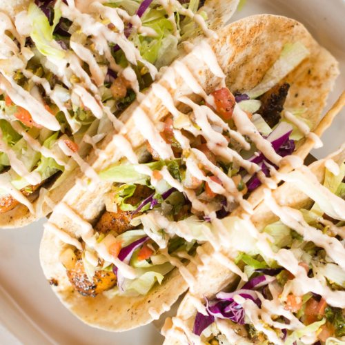 Four tacos filled with lettuce, tomatoes, and drizzled with sauce, served on a white plate.
