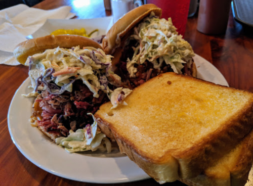 Two pulled pork sandwiches topped with coleslaw, served with a slice of toasted bread on a white plate.