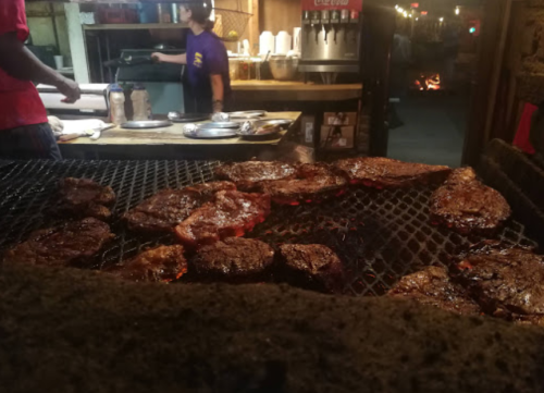 Grilling steaks on a barbecue with a person in the background preparing food in a lively outdoor setting.