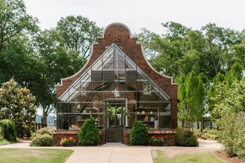 A brick greenhouse with large glass panels, surrounded by greenery and a pathway leading to its entrance.