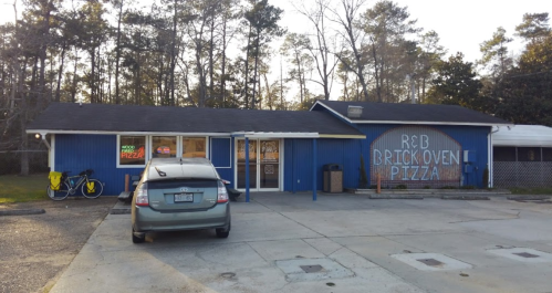 A blue building with a sign reading "R&B Brick Oven Pizza," featuring a parked car and a bicycle outside.