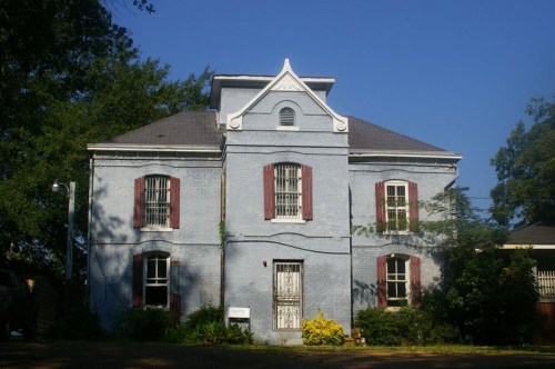 A two-story blue house with red shutters, surrounded by greenery and under a clear blue sky.