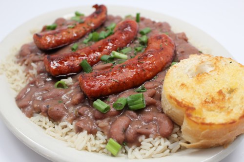 A plate of rice topped with red beans, grilled sausage, and garnished with green onions, alongside a slice of toasted bread.