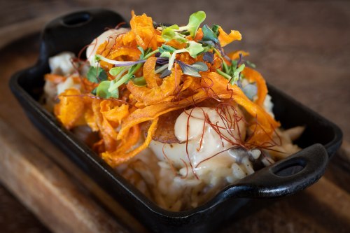 A close-up of a dish featuring creamy risotto topped with crispy sweet potato, microgreens, and saffron strands in a black bowl.