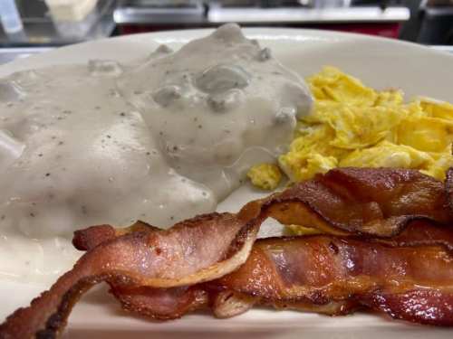 A plate of scrambled eggs, crispy bacon, and biscuits topped with creamy sausage gravy.