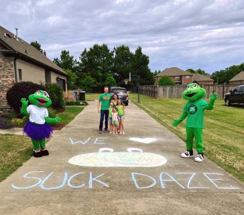 Two frog mascots stand on a driveway with a family, surrounded by grass and a cloudy sky, with "WE ❤️ SUCK DAZE" chalked on the ground.