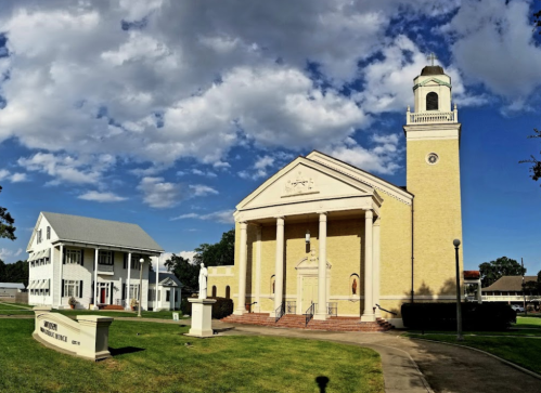 Historic yellow church with a clock tower, next to a white building, under a partly cloudy sky. Green lawn in foreground.