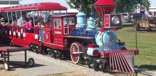 A colorful miniature train with passengers rides along a track in a sunny outdoor setting.