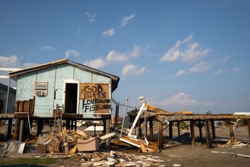 A damaged house on stilts with a sign reading "God Bless Louisiana" amidst debris and clear blue skies.