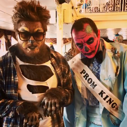 Two people in costumes: one as a werewolf with claws, the other as a zombie in a suit with a "Prom King" sash.