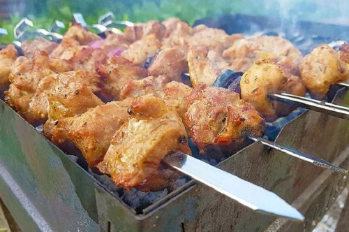 Grilled skewers of marinated chicken sizzling over charcoal, surrounded by smoke and greenery in the background.