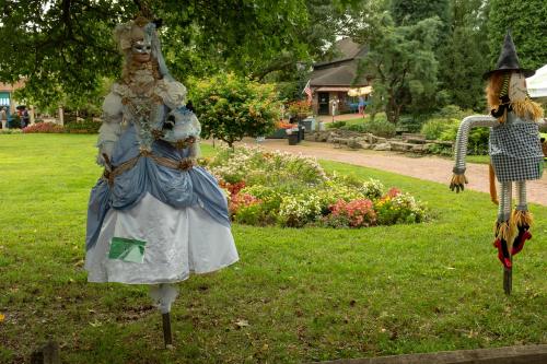 A whimsical scarecrow in a blue and white dress stands beside a traditional scarecrow in a garden with colorful flowers.
