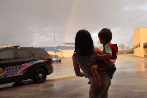 A child is held by an adult, both looking at a rainbow in the sky near a police vehicle.