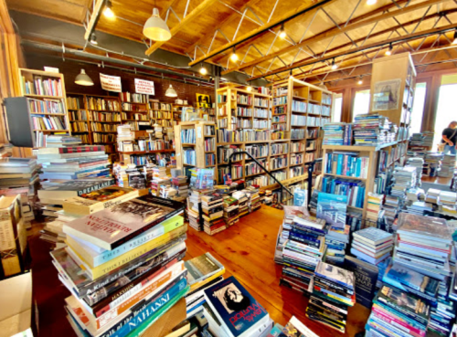 A cozy bookstore filled with tall shelves of books and stacks of books on the floor, creating a warm, inviting atmosphere.