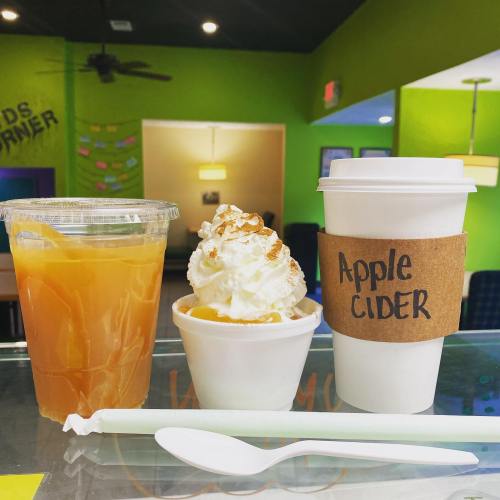 A clear cup of iced tea, a small dessert with whipped cream, and a cup of apple cider on a colorful café table.