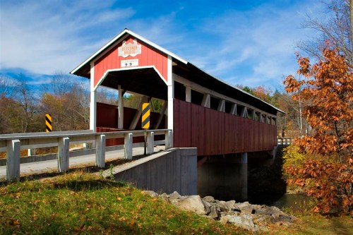 A red covered bridge spans a small stream, surrounded by autumn foliage and a clear blue sky.