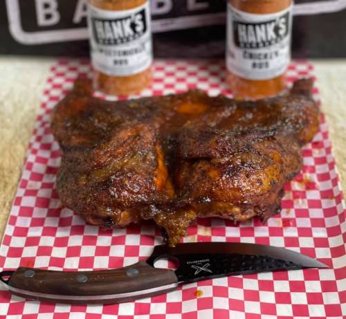 Grilled chicken coated in seasoning, displayed on a checkered paper with a knife and spice bottles in the background.