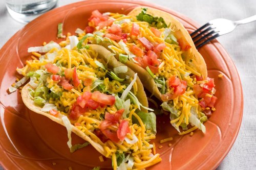 Two tacos filled with lettuce, tomatoes, and shredded cheese on an orange plate, with a fork and glass of water in the background.