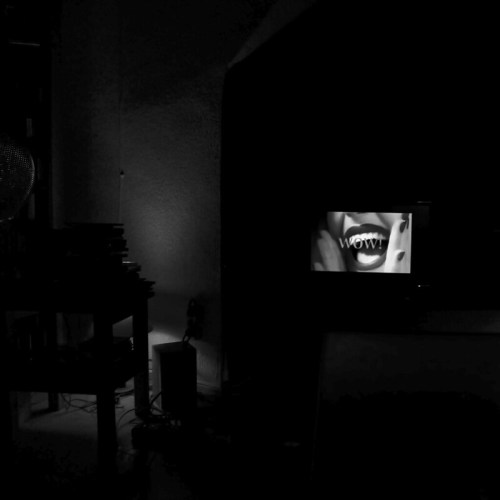 A dimly lit room featuring a TV displaying the word "WOW" with a close-up of a mouth, surrounded by shadows.