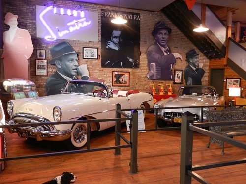A vintage car display featuring two classic cars, with large Frank Sinatra portraits and memorabilia in a cozy setting.
