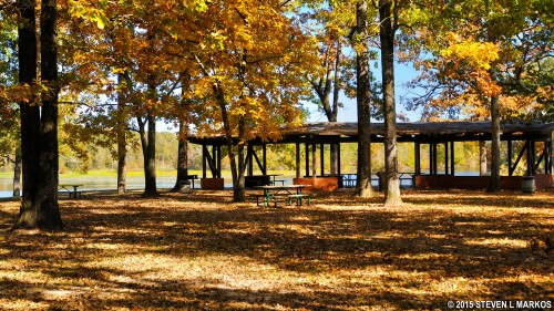A serene park scene with a pavilion surrounded by colorful autumn leaves and a calm lake in the background.